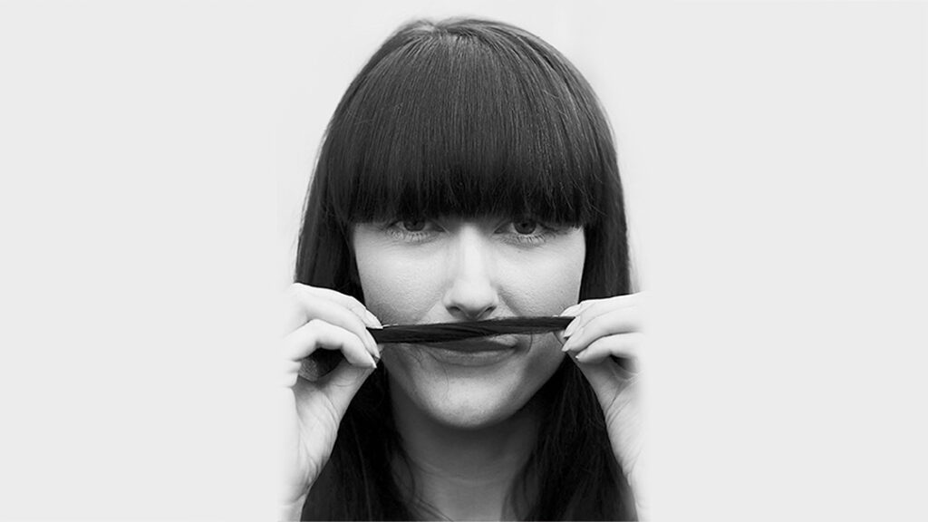 Femme brune à frange mimant une moustache avec une mèche de cheveux